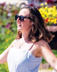 Bridget Anderson wearing sunglasses, opening her arms towards the left, with flowers in the background.