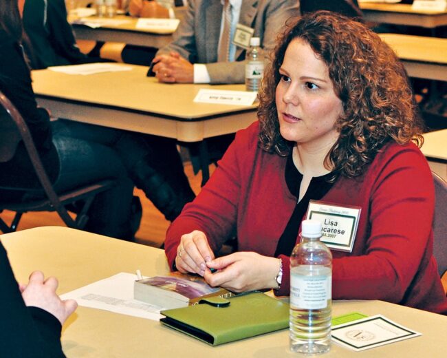 Woman in a red cardigan, sitting at a desk, listening to someone out of frame.