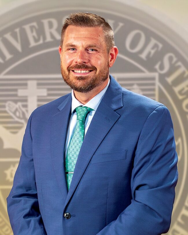 Chris Krivoniak wearing a suit and tie standing in front of a university seal.
