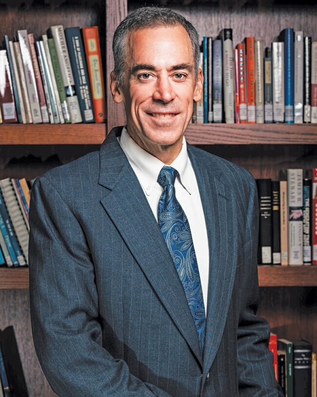 Dr. Ronald Bolster in a suit and tie, standing in front of a bookshelf.
