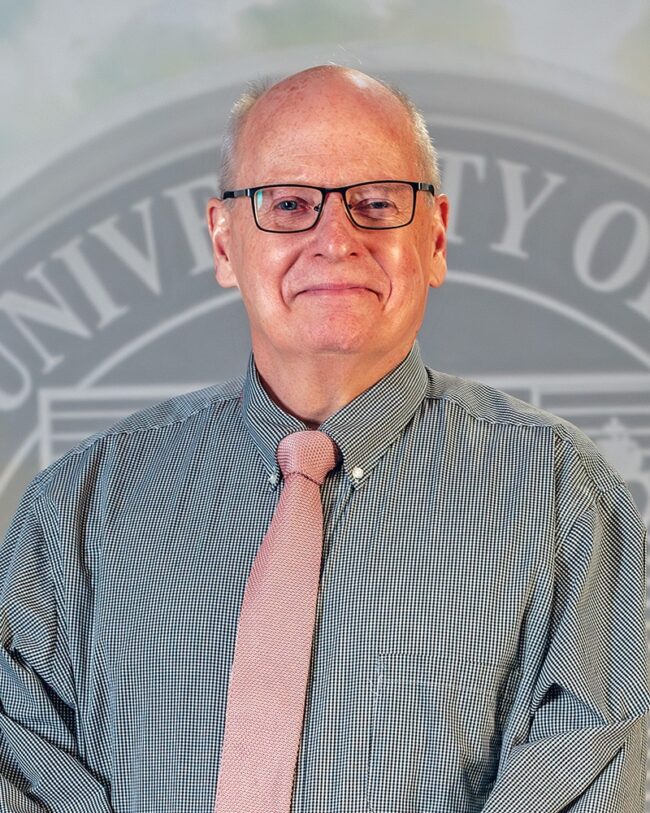 Dr. Timothy Williams wearing a gray button up and pink tie, standing in front of the university seal.
