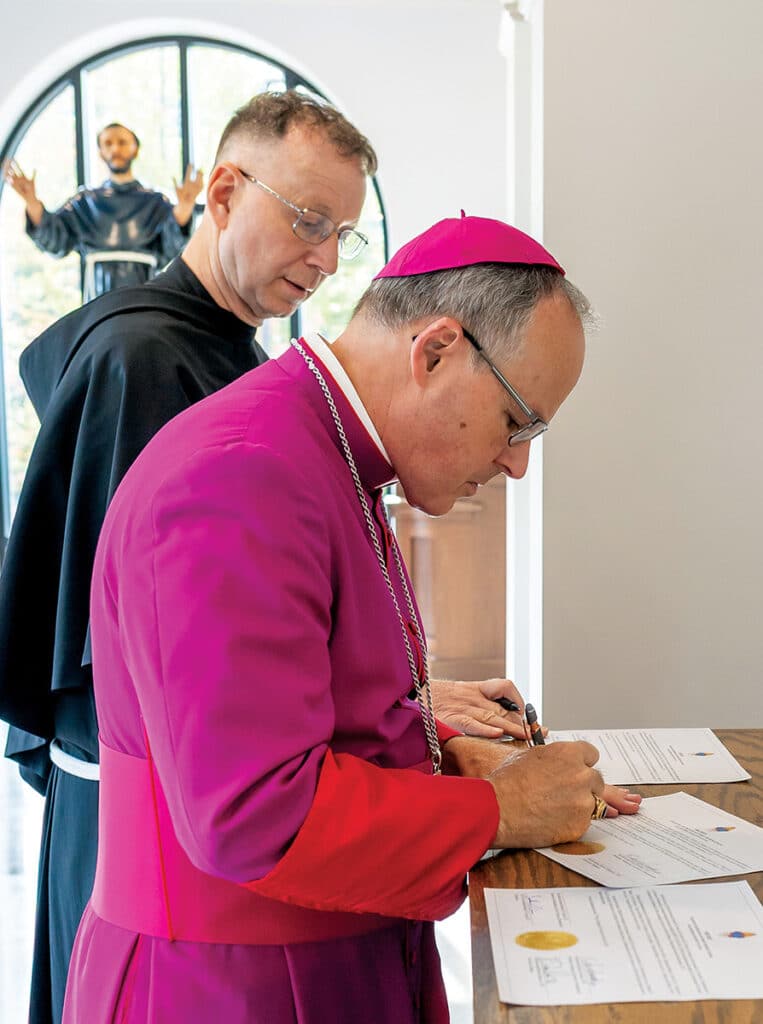 Fr. Shawn Roberson, TOR, watching as and Bishop Lohse signs papers. 