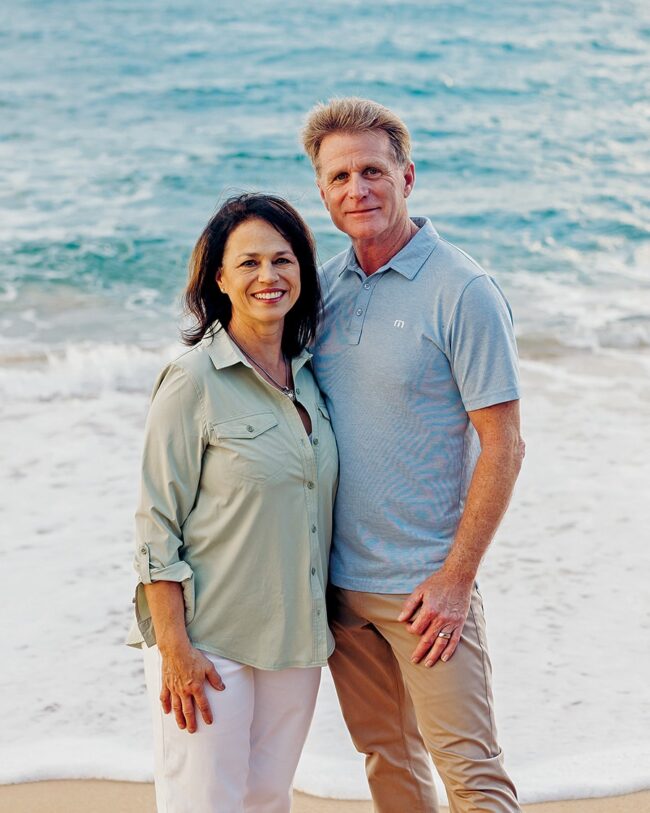 Greg and Martha McKenna smiling at the camera with a beach in the background.