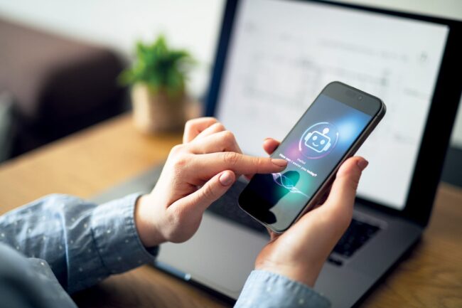 Two hands holding a phone with and image of a AI robot on the screen, with a desk in the background.