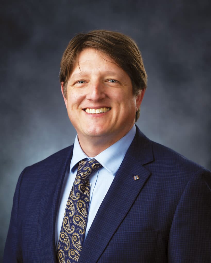 Patrick Flannery wearing a suit and tie in front of a gray background.