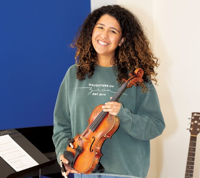 Alina Rodriguez holding a violin indoors, wearing a green sweatshirt, standing beside a piano and sheet music in a music practice space.