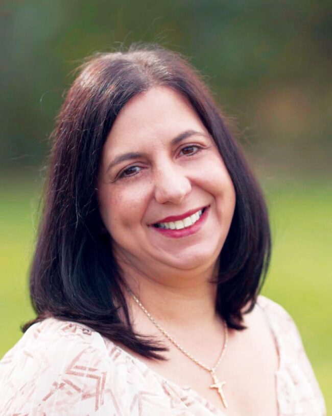 Caroline Gambale-Dirkes with dark hair wearing a light patterned blouse and cross necklace, photographed outdoors in a natural setting.