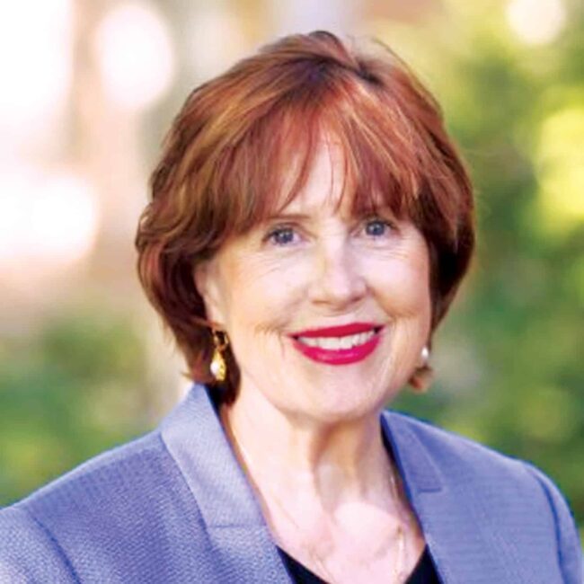Professional portrait of Dr. Deborah Savage with short auburn hair wearing a blazer and earrings, photographed outdoors against a softly blurred background.