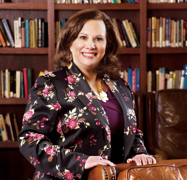 Dr. Regina Poletunow Boerio seated in a library, wearing a floral blazer and necklace, with bookshelves visible in the background.