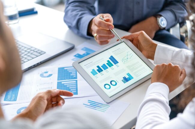 People reviewing financial charts and graphs on a tablet during a meeting, with printed reports and a laptop on the table.