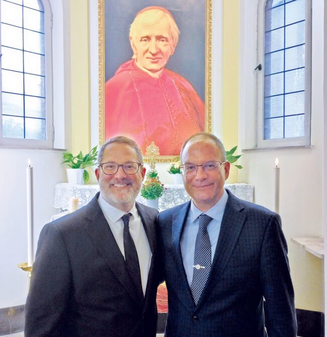 Dr. Logan Gage and coauthor Dr. Frederick Aquino standing together inside a chapel, with a framed portrait of a cardinal and lit candles visible behind them.