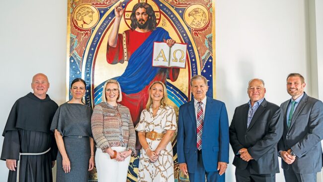Seven adults posing for a group photo in front of a large icon of Christ holding a book with the Alpha and Omega symbols.