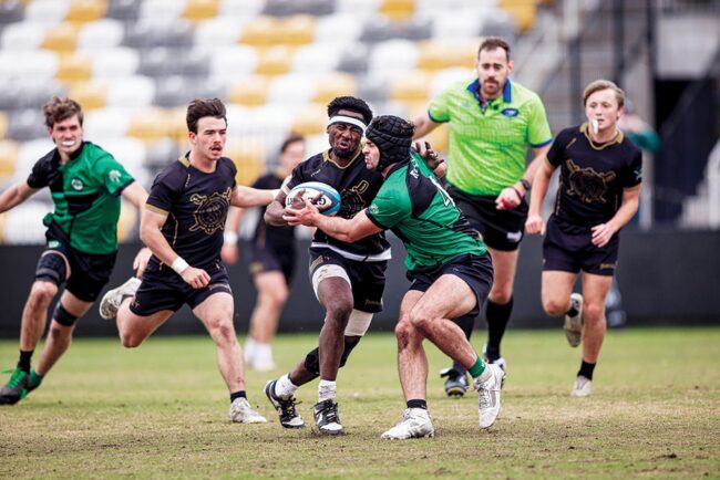 Rugby players in uniform competing during a match, with one player carrying the ball as opponents move in to tackle him on the field.