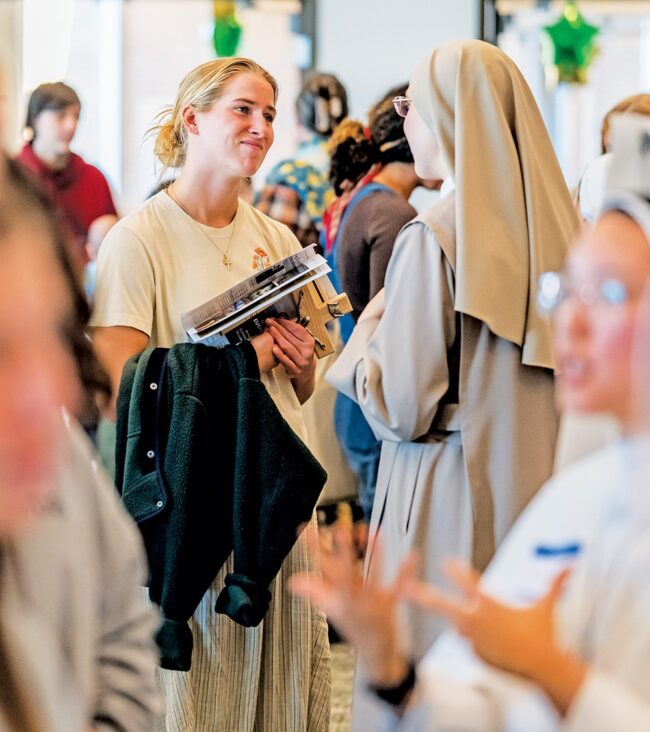 A student holding books talks with a woman in a religious habit at an indoor vocations fair with other attendees in the background.