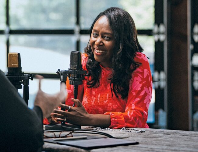 Immaculée Ilibagiza speaking into a podcast microphone during a recorded conversation, seated at a table in a studio environment.