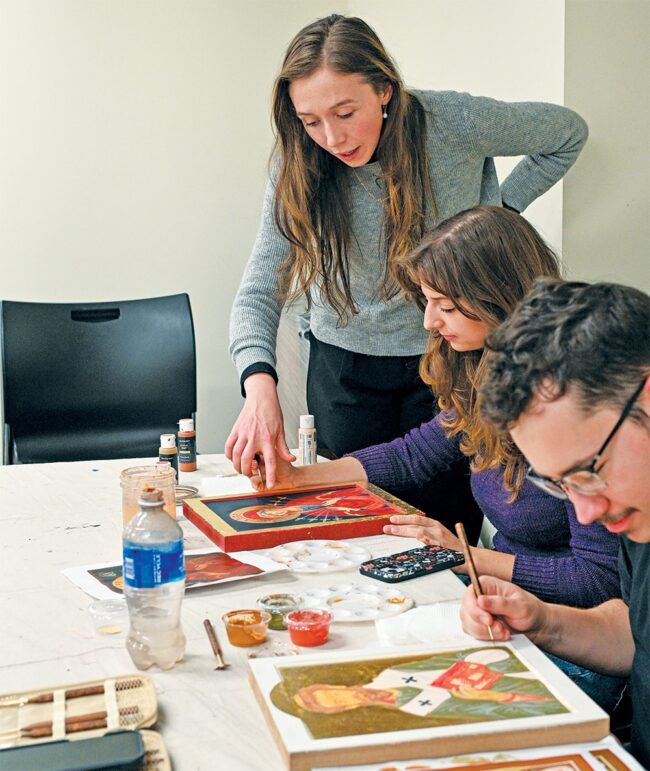 Students working together on religious icon painting in a studio setting, using brushes and paints on wooden panels as part of an art class.