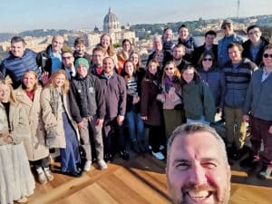 A large group of adults posing for a group selfie outdoors, with a city skyline and a domed building visible in the background.