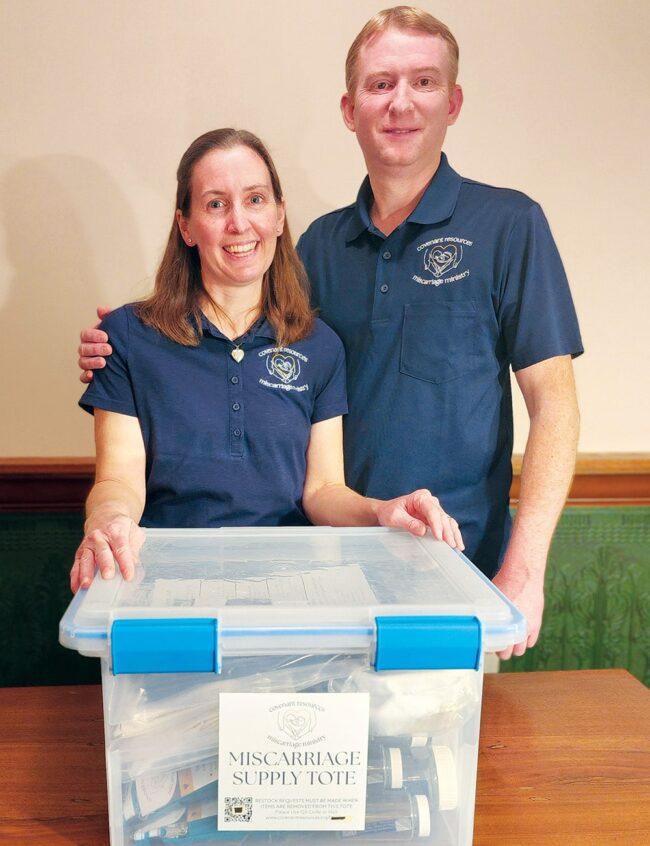 Rebecca and Chris ’98 Harpring standing indoors behind a clear container labeled “Miscarriage Supply Tote,” wearing matching Covenant Resources Miscarriage Ministry shirts.