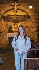 Young woman standing inside a stone chapel, holding a rosary, with an altar and crucifix visible behind her.