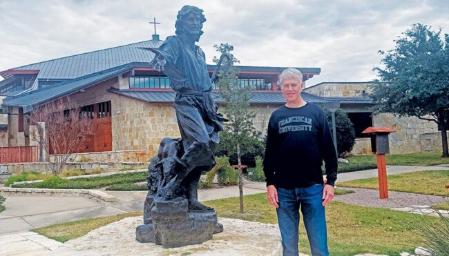 Man standing outdoors next to a bronze statue of a robed figure with outstretched arm, with a stone building visible in the background.