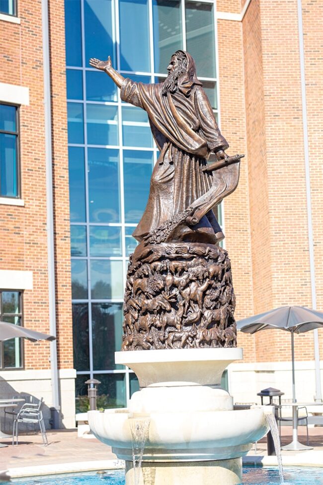 Bronze statue of Noah standing atop a fountain, with one arm extended and a scroll in his hand, set in front of a brick and glass building.