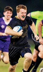 Brendan Lacey breaking away from Holy Cross defenders during a rugby match under stadium lights.
