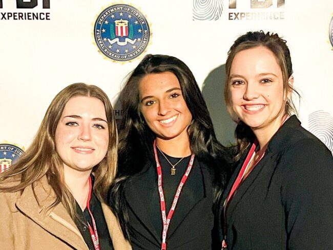 Three women posing in front of an “FBI Experience” backdrop while wearing event badges.