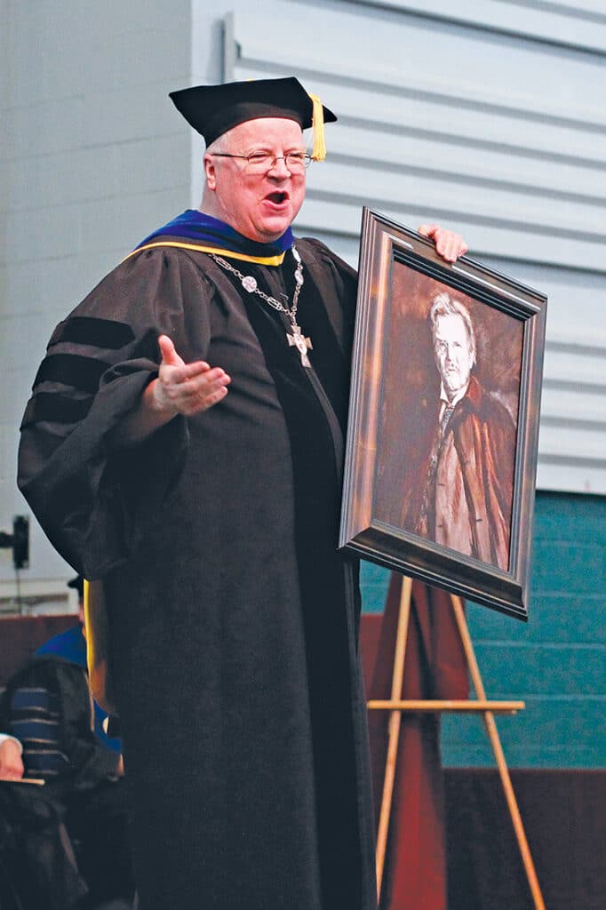 Fr. Terence Henry, TOR, speaking at a commencement ceremony while holding a framed portrait.