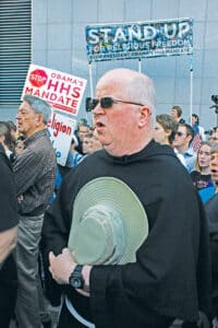 A Franciscan friar standing in a crowd at a rally with signs reading “Stand Up for Religious Freedom” and “Stop Obama’s HHS Mandate.”