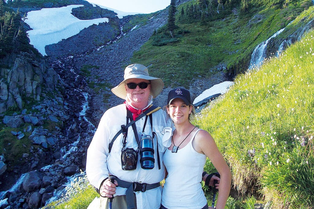 Fr. Terry and Kaitlyn Koehr Brindle hiking in a mountain landscape with waterfalls and snow-covered slopes.