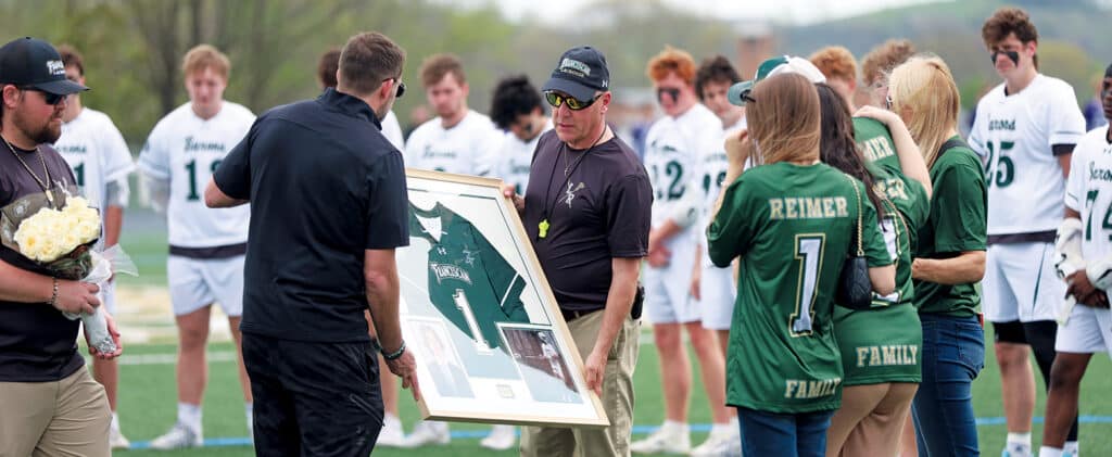 Franciscan lacrosse team and family members honoring Luke Reimer with a framed jersey presentation on the field.