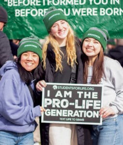 Three women wearing Franciscan hats holding a sign that reads “I Am the Pro-Life Generation” at an outdoor event.
