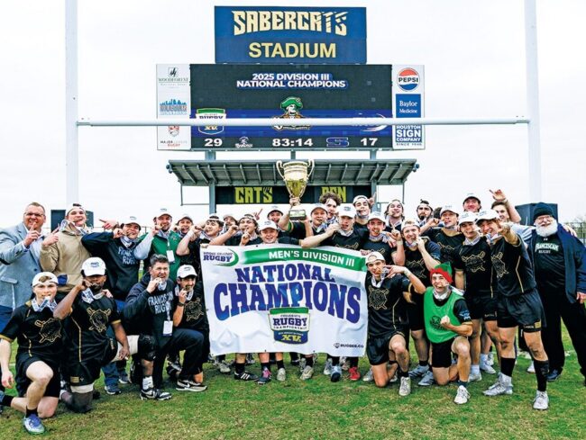 Rugby players and coaches posing on a field with a “National Champions” banner and trophy beneath a stadium scoreboard.