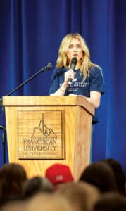 Riley Gaines speaking at a Franciscan University of Steubenville event from a podium on stage.