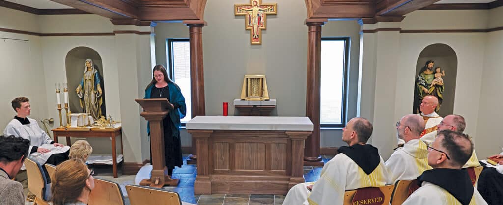 People seated in a chapel as a woman reads from a lectern near the altar.