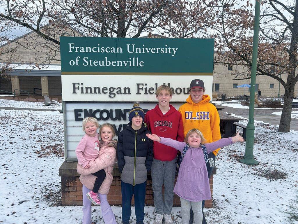 Six children standing in front of a sign reading “Franciscan University of Steubenville” and “Finnegan Fieldhouse.”