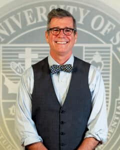 Portrait of Dr. Bob Rice wearing glasses, a bow tie, and a vest in front of the university seal.