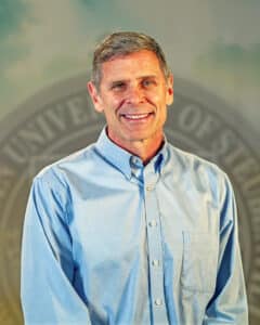 Portrait of Dr. Jeffrey Rohde wearing a light blue button-down shirt in front of the university seal.
