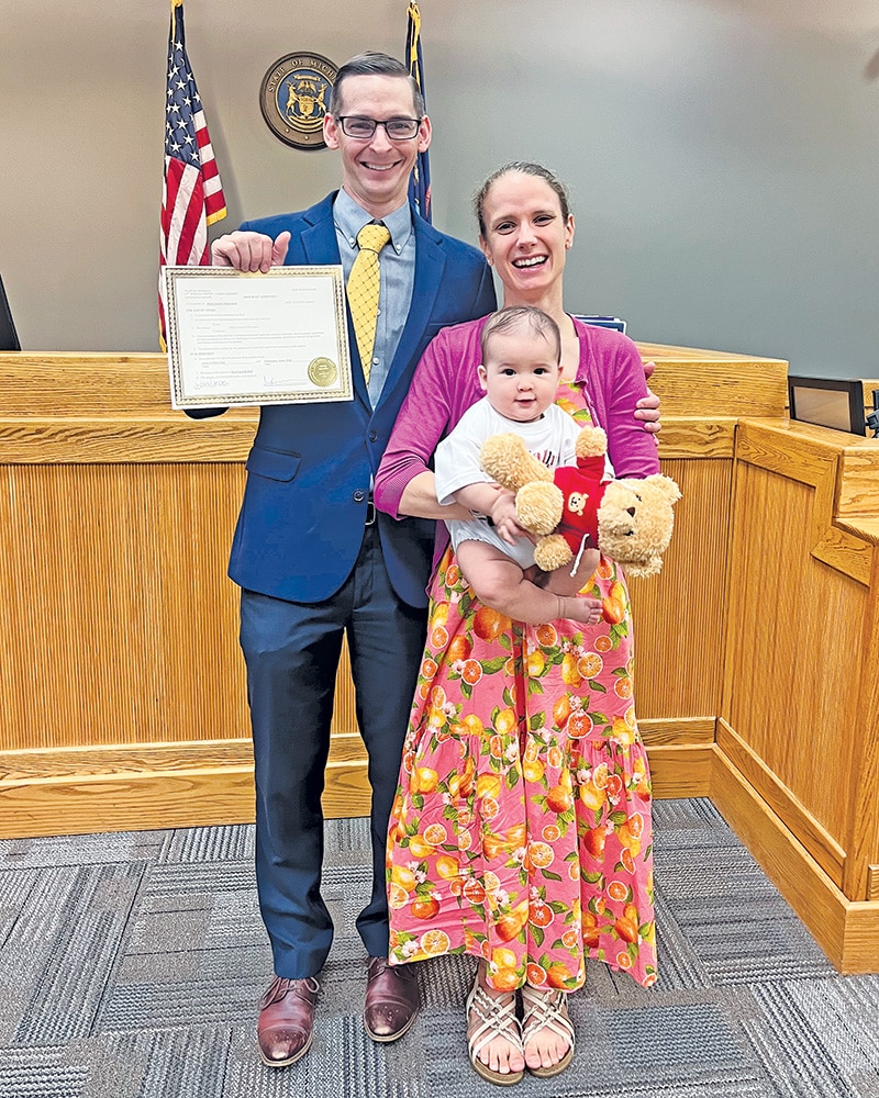 A man, Chris Hoff ’14 MA ’16, holding a certificate stands with a woman, Isabel (Peck ’15) Hoff and baby in a courtroom with U.S. and Michigan flags behind them.