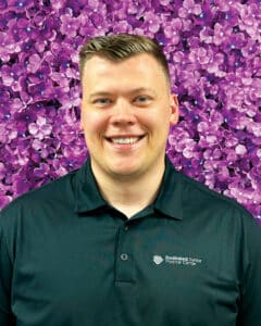 Portrait of a Joseph Rall ’18, DO wearing a branded polo in front of purple flowers.