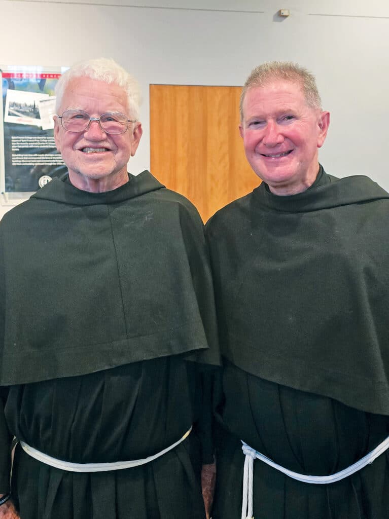 Two Franciscan friars standing together in traditional habits with white cord belts.