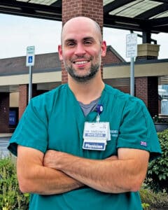 Physician in green scrubs wearing a “Dr. Sweeney, Physician” ID badge outside a medical facility.