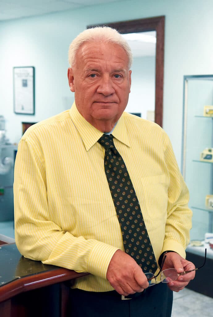 Portrait of Ron Vondenichar standing indoors in a yellow dress shirt and tie, holding his glasses.